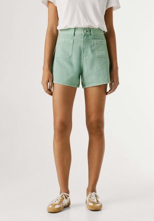 Frau trägt mintgrüne Shorts mit hohem Bund und Fronttaschen, weißes T-Shirt und beige Sneaker mit weißen Schnürsenkeln, steht vor einfarbigem Hintergrund.