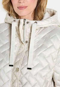 Gepolsterte, hellfarbige Jacke mit Kapuze. Verfügt über silberne Hardware, einen Front-Reißverschluss und weiße Kordeln mit Branding. Weicher, strukturierter Stoff.