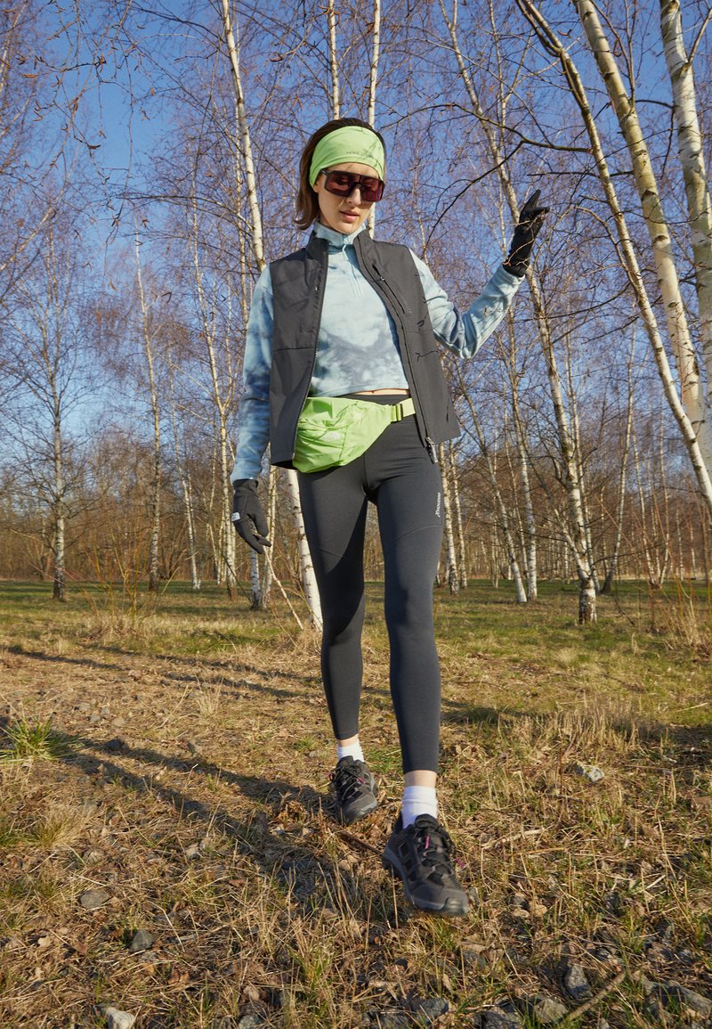 Kvinna som bär solglasögon, grön hårband, väst och leggings, går utomhus bland björkar en solig dag.