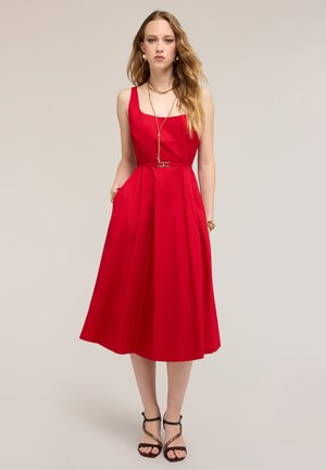 Kobieta stojąca, ubrana w czerwoną midi sukienkę bez rękawów z kieszeniami, uzupełnioną warstwowymi naszyjnikami, bransoletkami i czarnymi sandałami na obcasie z paskami.