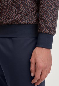 Sweatshirt navy avec motif géométrique orange, avec des poignets et un ourlet côtelés, présentant une texture douce. Main posée sur le tissu navy en dessous.