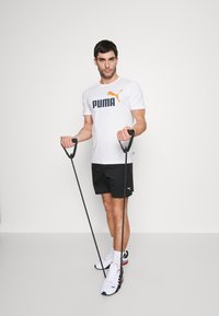 Balta trumpa rankovėmis marškinėliai su juodu ir oranžiniu "PUMA" logotipu, juodos šortai ir balti batai su raudonais akcentais; laiko treniruočių juostas.