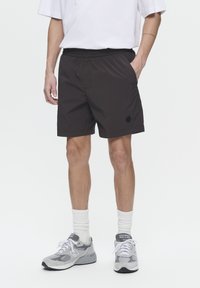 Shorts de sport noirs avec une taille élastique, dotés de poches latérales et d'un petit logo noir. Portés avec des baskets grises et des chaussettes blanches.