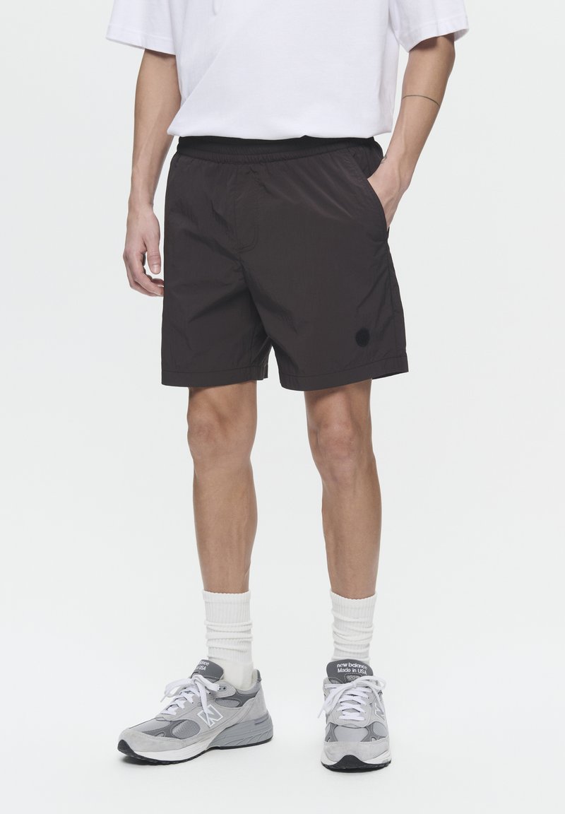 Shorts de sport noirs avec une taille élastique, dotés de poches latérales et d'un petit logo noir. Portés avec des baskets grises et des chaussettes blanches.