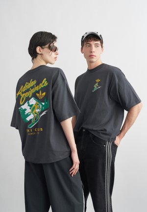 Dvaja mladí dospelí v tmavých tričkách Adidas Originals s grafikou lyžovania, stoja na jednoduchom svetlosivom pozadí.