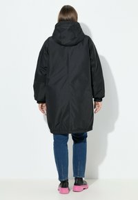 Zwarte waterdichte parka met een capuchon, elastische manchetten en een getailleerde zoom. Draagt over een blauwe jeans en schoenen met een roze zool.