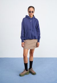 Sweatshirt azul-marinho com cordões e bolso frontal, combinado com uma saia mini bege com botões. Ténis castanhos e verdes complementam o visual.