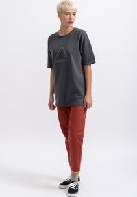 Grijze oversized t-shirt met een driehoekige grafische print, gecombineerd met roestkleurige joggers. Zwarte sneakers en contrasterende gele accenten op de sokken.