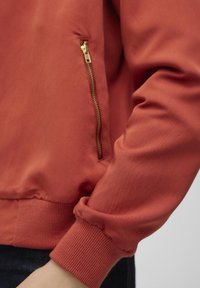 Veste bomber rouge avec une finition lisse, des poignets côtelés et une poche latérale zippée avec des éléments en métal doré.
