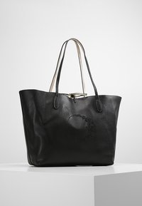 Musta nahkainen olkalaukku, jossa on tekstuuri, pitkät kahvat ja kohokuvioitu pyöreä kuvio etupuolella. Laaja avaus, minimalistinen muotoilu.