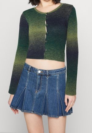 Kobieta ubrana w krótki, zapinany na guziki sweter z długim rękawem w zielonych odcieniach gradientu oraz niebieską plisowaną mini spódnicę z denimu.