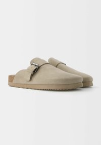 Sabots en Daim Beige avec un orteil arrondi, une sangle à boucle réglable et une semelle en liège. Semelle texturée avec un motif en zigzag pour l'adhérence.