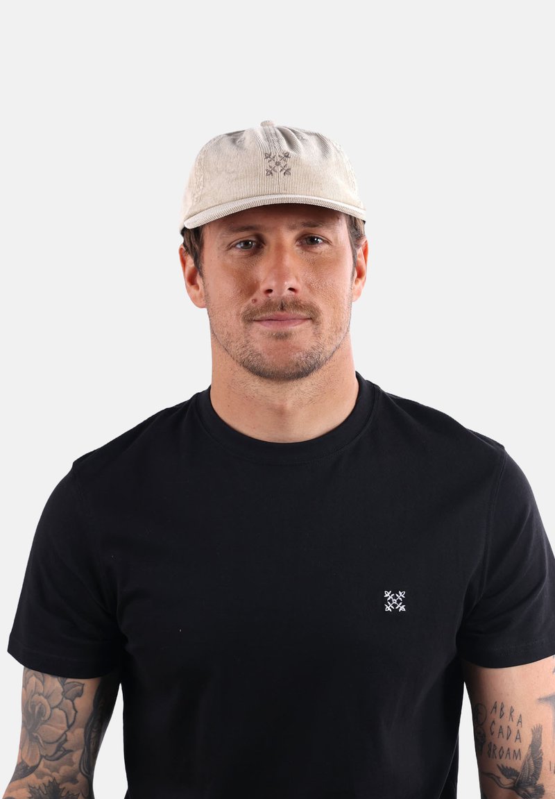 Casquette en velours côtelé beige clair avec une visière courbée et un design brodé. Portée avec un t-shirt noir arborant un petit logo blanc.