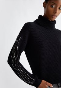 Maglione a collo alto nero in maglia a coste con applicazioni di borchie argentate lungo le maniche, caratterizzato da una vestibilità rilassata e una texture morbida.