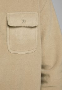 Beżowa kurtka z polaru o strukturalnej powierzchni, z patką z kieszenią zapinaną na guzik na lewej części klatki piersiowej. Miękki i ciepły materiał.