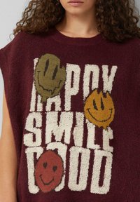 Weinroter Pullover mit strukturiertem weißen Text, der "HAPPY SMILE GOOD" liest, und drei bunten Smileys in Olivgrün, Orange und Rot.