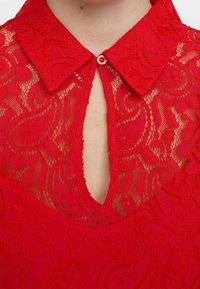 Red lace blouse featuring a collar and a keyhole opening with a button detail. The texture is intricate with floral patterns throughout.