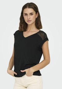 Czarny t-shirt z krótkim rękawem i dekoltem w serek, z koronkowymi akcentami na ramionach i luźnym krojem. Materiał wydaje się miękki i lekki.