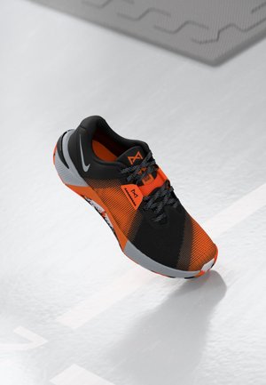Chaussure de sport noire et orange avec des lacets gris à motifs et une semelle blanche, flottant au-dessus d’un sol texturé gris clair avec des marques géométriques.