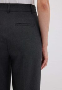 Lato destro di una persona che indossa pantaloni grigi scuri su misura con una tasca a filetto sul retro e una maglietta bianca infilata dentro.