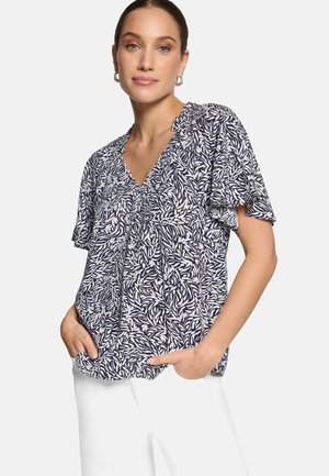 Femme portant un chemisier à manches courtes bleu marine et blanc à motif de feuilles avec un col en V, et un pantalon blanc, les mains dans les poches, sur un fond uni.