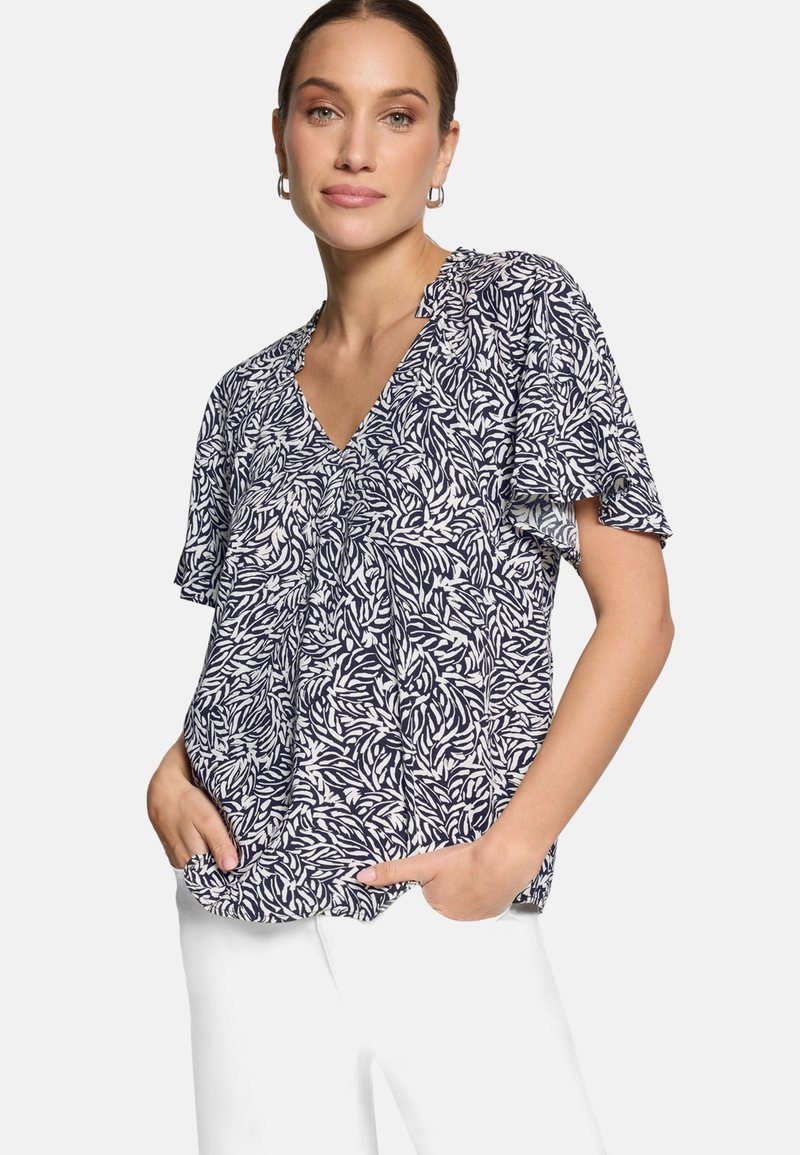 Femme portant un chemisier à manches courtes bleu marine et blanc à motif de feuilles avec un col en V, et un pantalon blanc, les mains dans les poches, sur un fond uni.