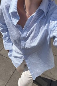 Light blue striped button-up shirt with textured fabric, featuring a relaxed fit, oversized collar, and long sleeves. Worn over cream pants.