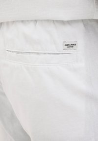 Fehér vászon nadrág, hátulján egy téglalap alakú címkével ellátott zseb található, amelyen a "JACK & JONES" felirat olvasható, és a zseb szélén keskeny varrás látható.