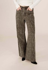 Personne portant un pantalon large à taille haute en imprimé léopard, les mains dans les poches et des chaussures noires à talons pointus, devant un fond uni.