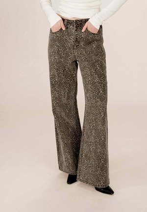 Personne portant un pantalon large à taille haute en imprimé léopard, les mains dans les poches et des chaussures noires à talons pointus, devant un fond uni.