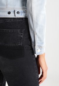 Hellblaue Jeansjacke mit schwarzen Knöpfen, kombiniert mit schwarzen Jeans mit strukturierter Oberfläche und einer Gesäßtasche. Sichtbarer silberner Logo-Akzent.