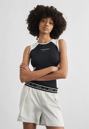 Frau mit lockigem Haar, die ein schwarzes und weißes ärmelloses Nike Shox Top und helle Shorts mit Nike-Logo am Bund trägt, steht mit verschränkten Armen.