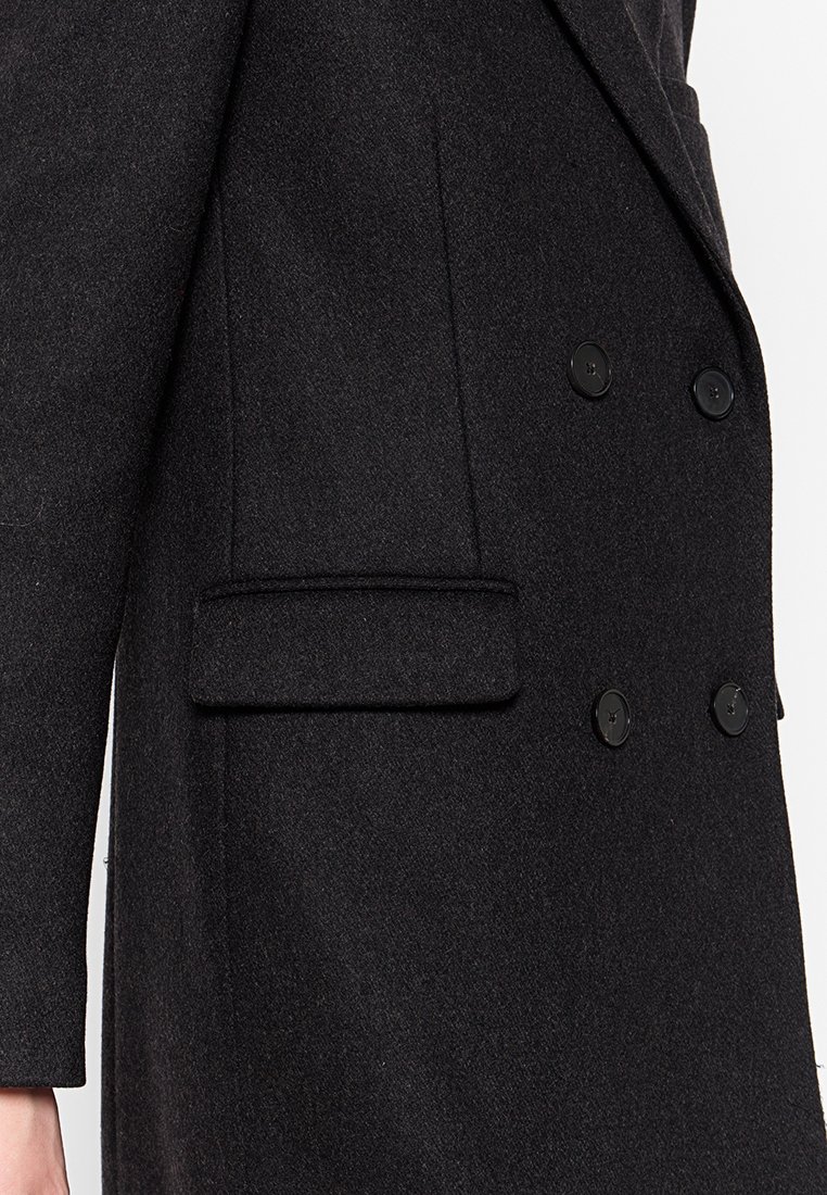 Manteau noir en laine à double boutonnage avec un col cranté, tissu texturé, poche latérale unique et boutons ronds noirs.