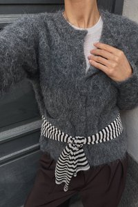 Cárdigan gris esponjoso con un escote redondo, detalles de botones y un cinturón atado de rayas en blanco y negro. Pantalones marrón oscuro visibles abajo.