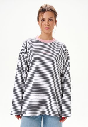 Vrouw draagt een losse zwart-wit gestreepte lange mouw blouse met een roze kraag en lichtblauwe spijkerbroek, staand tegen een witte achtergrond.