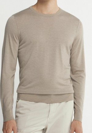 Homme portant un pull beige à col rond et manches longues ainsi qu'un pantalon clair, debout devant un fond uni clair.