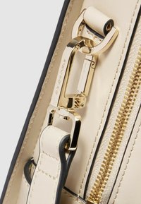 Detail van een beige leren tas met gouden hardware, een stevige clip en een soepele ritssluiting met een contrasterende zwarte rand. Zichtbare gestructureerde stiksel.
