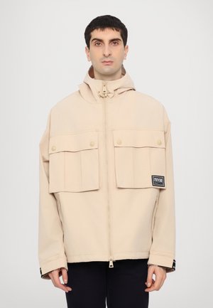 Veste à capuche beige en matériau léger, avec de grandes poches avant, une fermeture éclair et un patch logo sur la manche gauche.