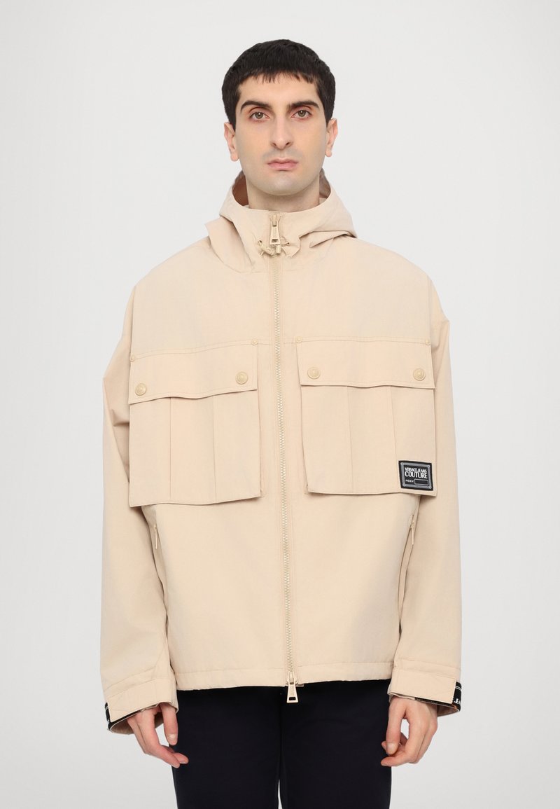 Beige hooded jacket made of lightweight material, featuring large front pockets, a zip closure, and logo patch on the left sleeve.