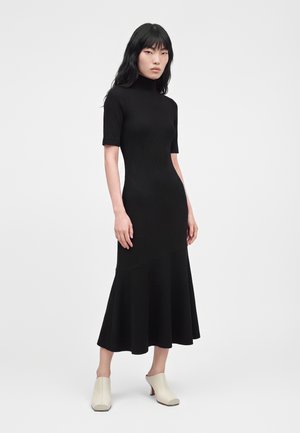 Femme aux longs cheveux noirs porte une robe midi noire ajustée à manches courtes et des chaussures crème à talons blocs, debout devant un fond uni.