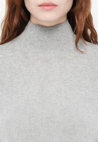Siv turtleneck pulover, izdelan iz mehkega pletenega materiala s tesnim krojem. Ima rebrasto teksturo na ovratniku in gladko telo.