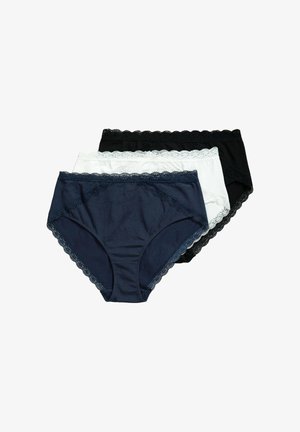 Femme portant un bralette en dentelle noire et une culotte taille haute assortie, debout contre un mur blanc, main posée sur la hanche.