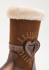Bottine en suède brun avec un revers en peluche beige, accents en bronze, boucle en forme de cœur et motif décoratif de points dorés.