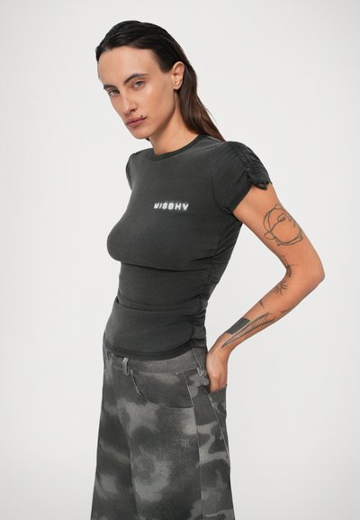 Femme aux cheveux bruns portant un t-shirt noir ajusté "MISBHV" et un pantalon camouflage gris, affichant des tatouages sur son bras droit, posant devant un fond blanc.