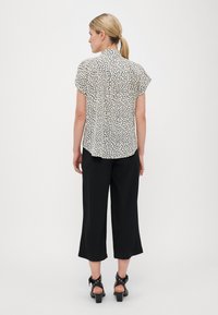 Blouse blanche à manches courtes avec un motif de pois abstraits noirs, avec un dos boutonné, associée à des culottes noires et des sandales à talons noires.
