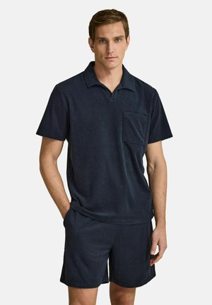 Man die een donkerblauw poloshirt met korte mouwen en borstzak draagt, gecombineerd met bijpassende korte broek, staand met één hand in de zak tegen een effen achtergrond