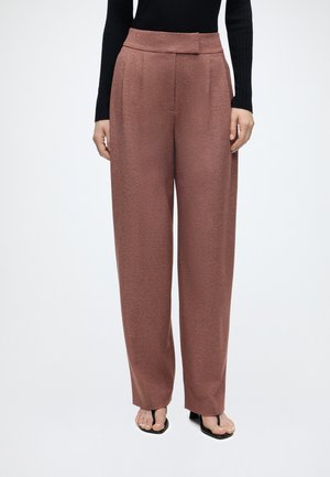 Femme portant un pantalon marron taille haute à jambes larges, un haut noir à manches longues et des sandales noires à bout ouvert sur un fond uni.