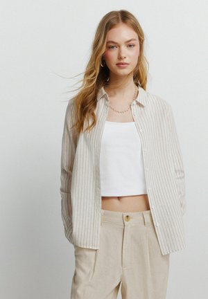 Młoda kobieta z długimi blond włosami, ubrana w beżową koszulę w paski zapinaną na guziki, na białym krótkim topie oraz beżowe spodnie z wysokim stanem, na tle jednolitego tła.