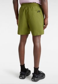 Persona con pantalones cortos color verde oliva que tienen un bolsillo trasero con cremallera, calcetines negros y zapatillas negras, de pie contra un fondo blanco.