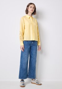 Camicia gialla a maniche lunghe con motivi a disegno sulle maniche, abbinata a jeans a gamba larga blu e sneakers metalliche argento.
